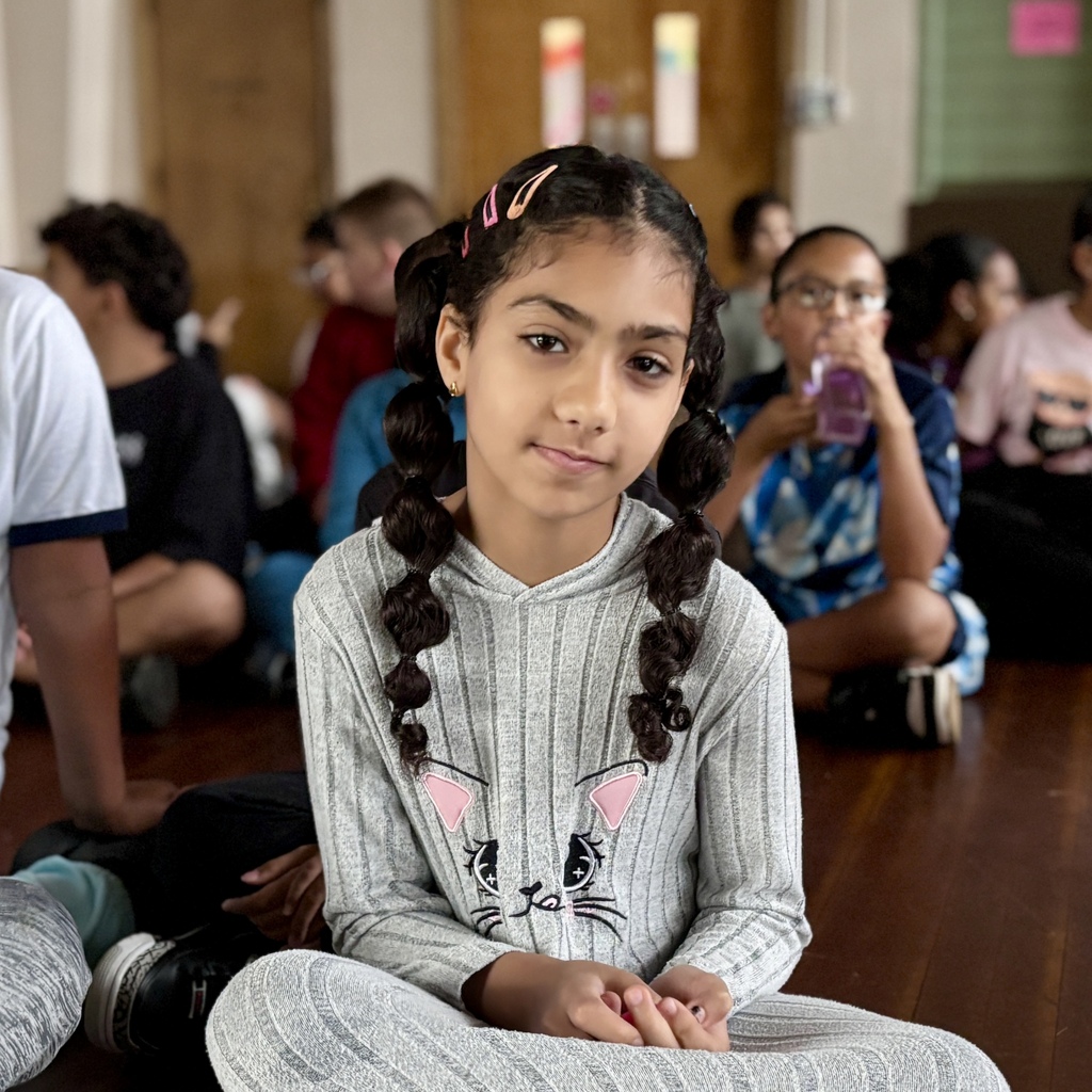 A young girl with braided hair and hair clips sits cross-legged on a wooden floor in a classroom.