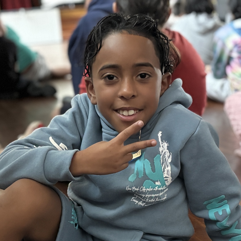 A smiling young boy in a blue hoodie makes a peace sign with his hand.