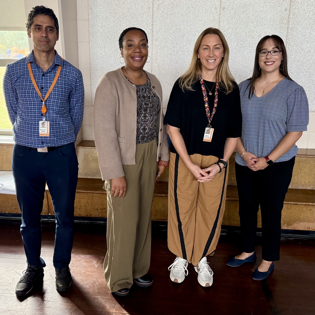Four teachers stand side-by-side indoors, smiling at the camera, wearing professional clothing and ID lanyards.