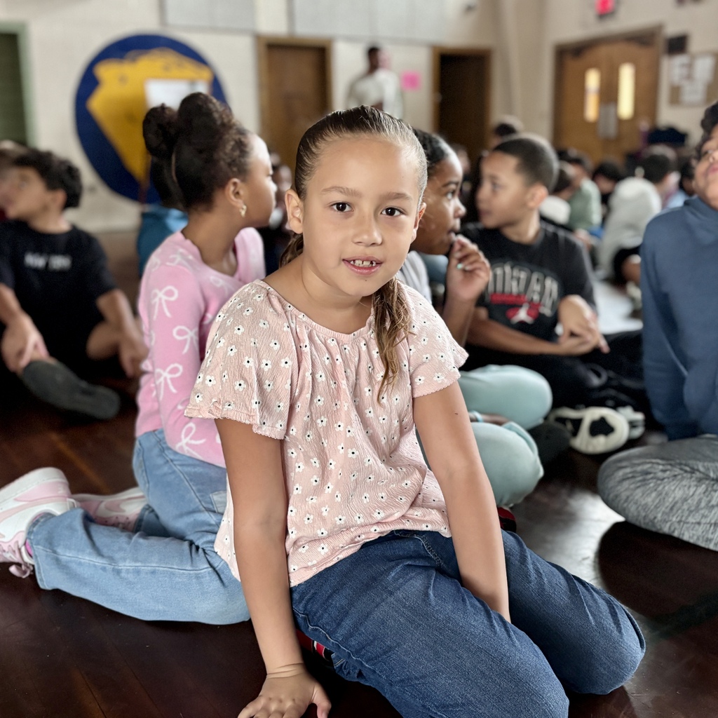 A young girl in a pink floral shirt looks forward while sitting on a wooden floor among other children