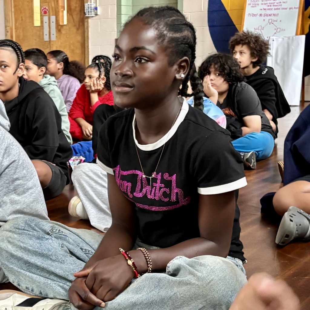 A young girl with braided hair sits on a wooden floor, looking attentively forward among a group of peers.