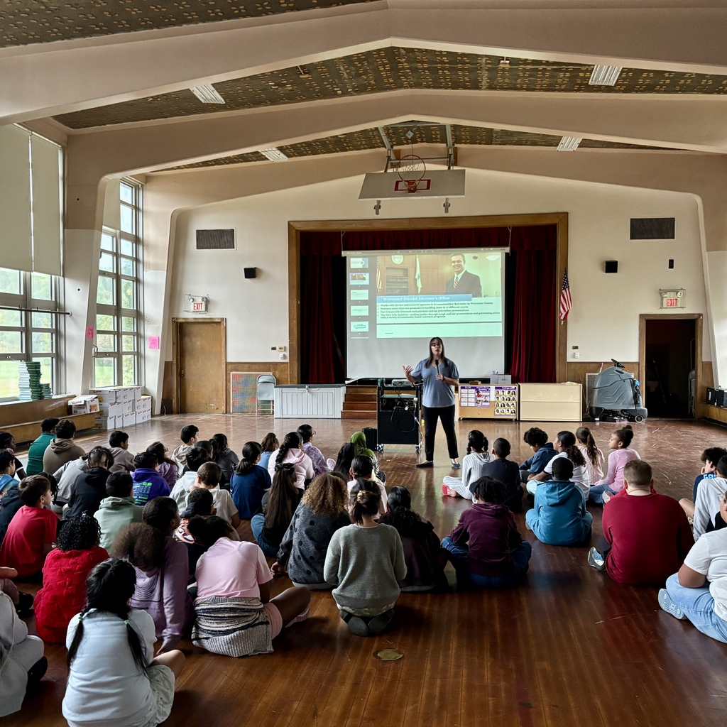 A woman gives a presentation to a large group of seated children in a gymnasium.