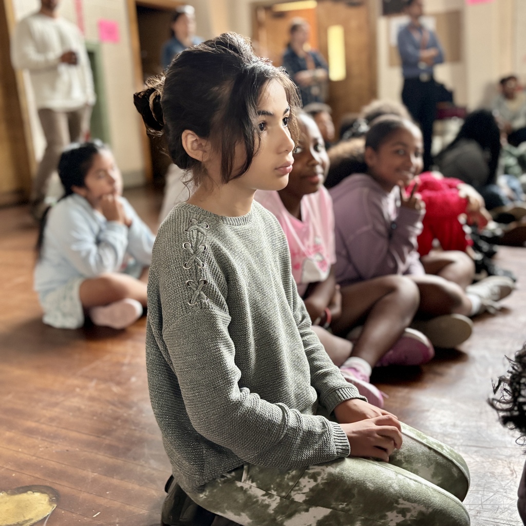 A young child in a grey sweater sits attentively on a wooden floor among peers.