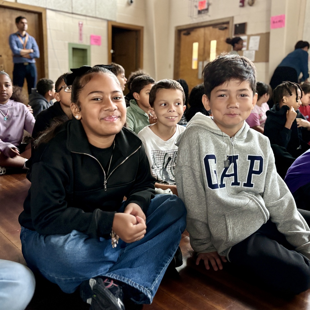 A group of children sit together on a wooden floor in a gymnasium or school room, smiling towards the camera.