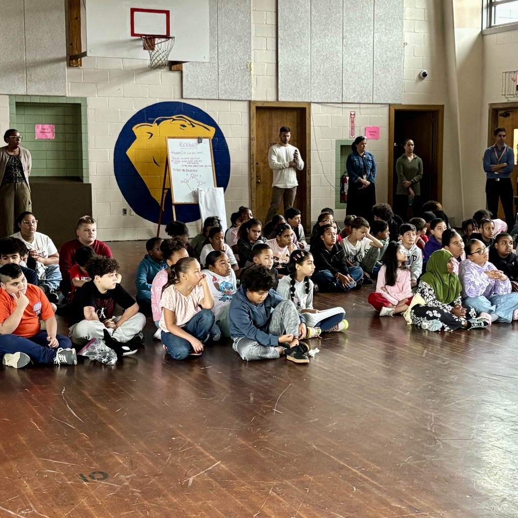 A group of children sit attentively on a gym floor during an indoor assembly.