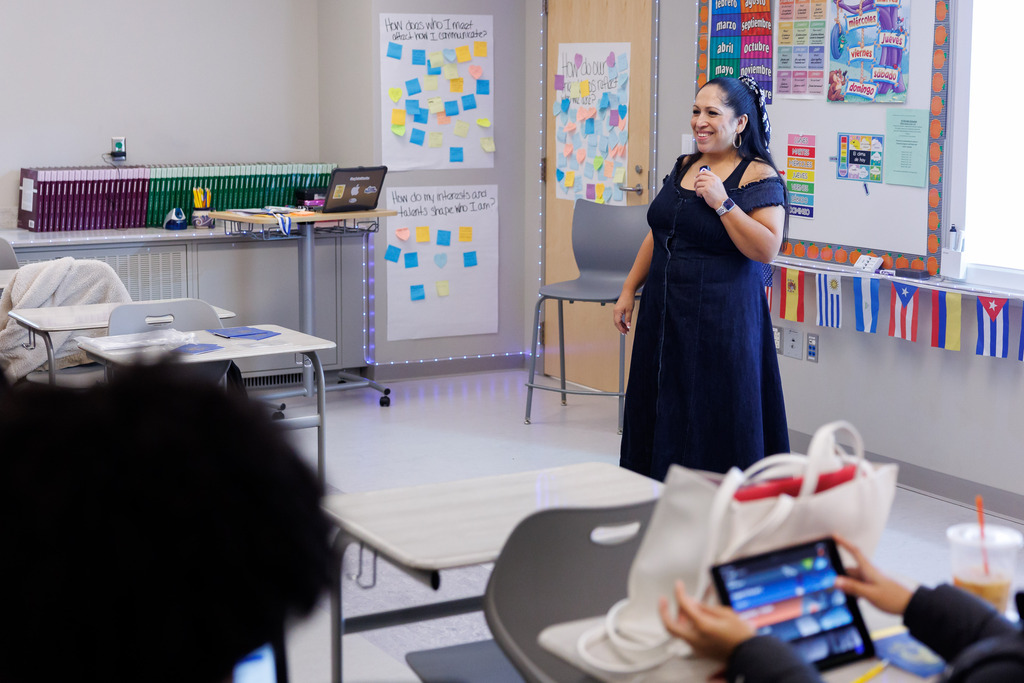 A teacher leads the classroom while standing at the front of the room and smiling.