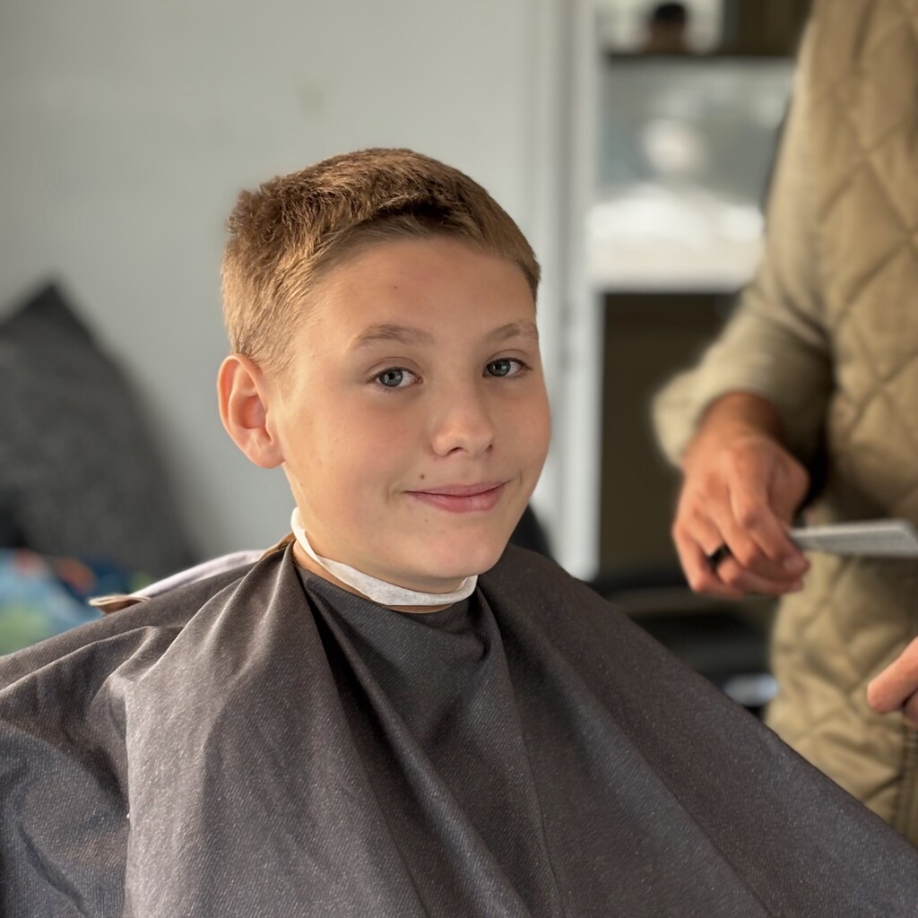 A young boy with short brown hair smiles while sitting in a salon chair wearing a cape.