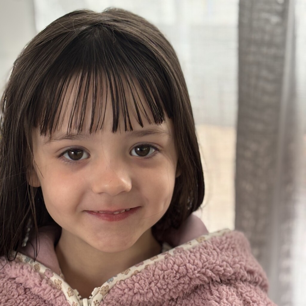 A close-up portrait of a young child with dark hair and bangs, smiling gently at the camera.