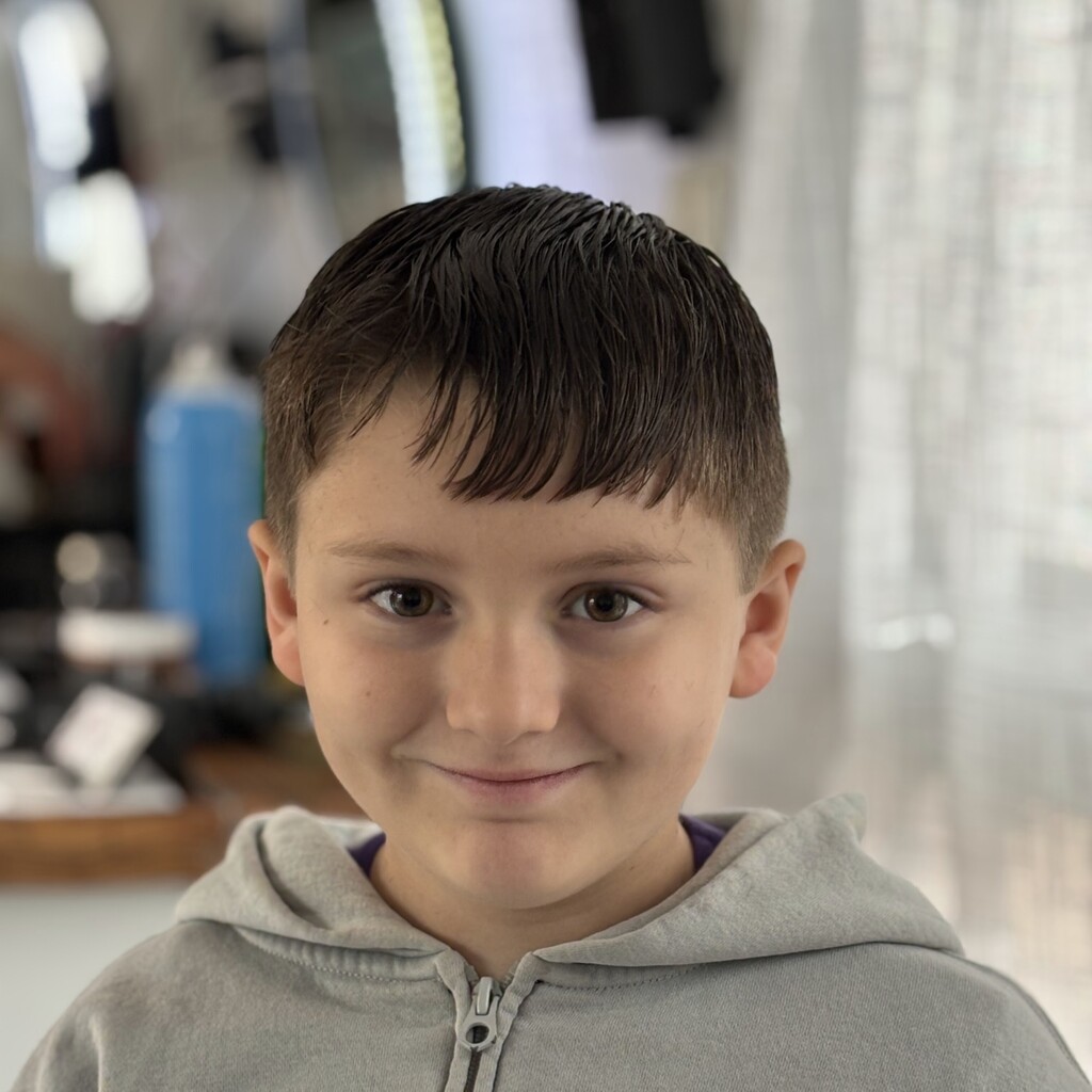 A young person with short brown hair and a gentle smile wears a gray zip-up hoodie indoors.