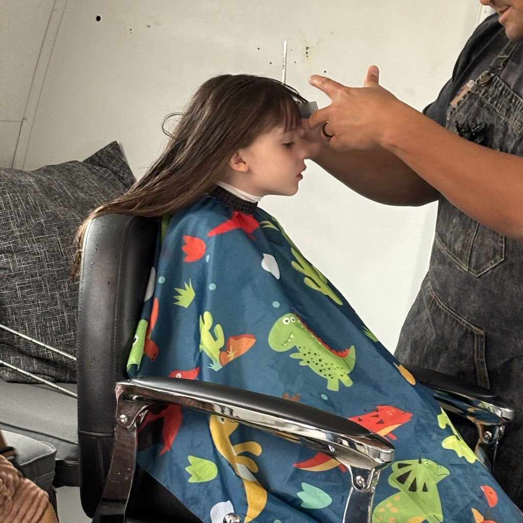 A child sits calmly in a salon chair wearing a blue dinosaur print cape while getting bangs trimmed.