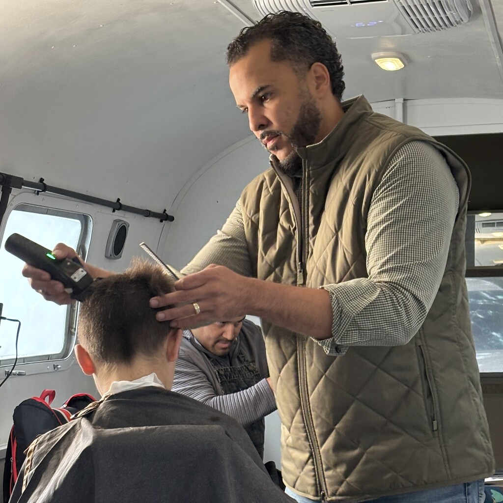 A stylist wearing a quilted vest uses clippers and a comb to cut a client's hair.