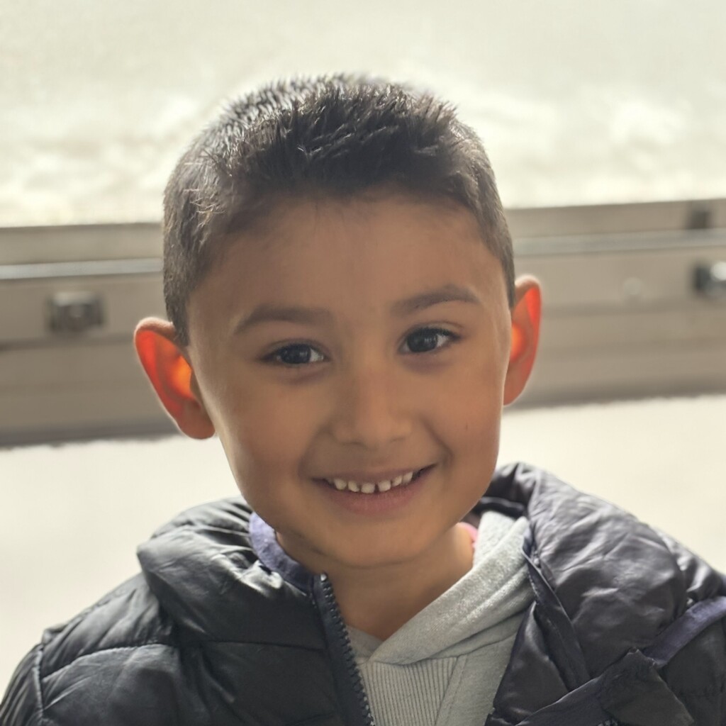 A young child smiles warmly at the camera, wearing a dark puffer jacket over a grey hoodie.