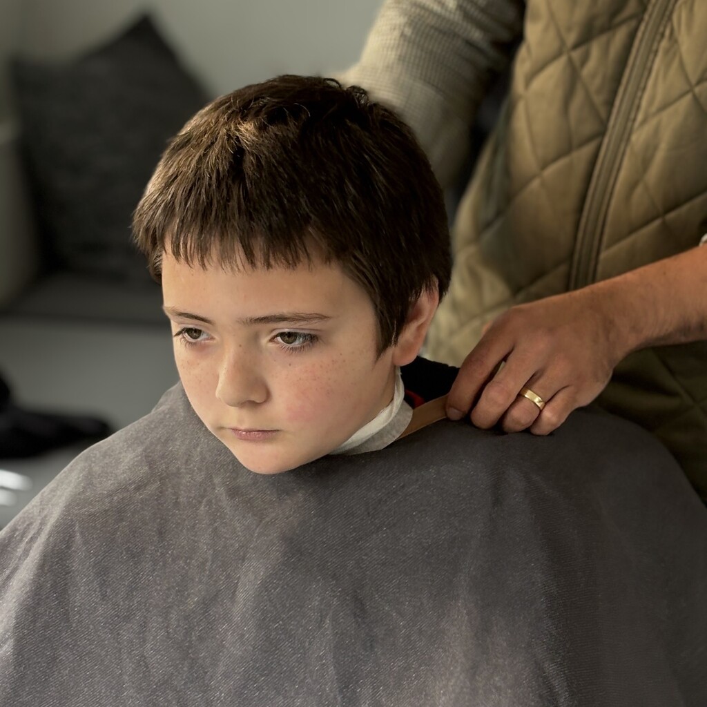 A young person sits quietly while wearing a grey hairdressing cape indoors.