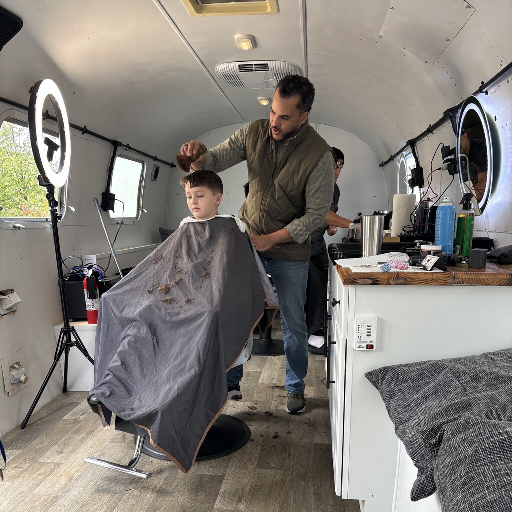 A stylist brushes hair from a young boy's cape inside a mobile hair salon van.