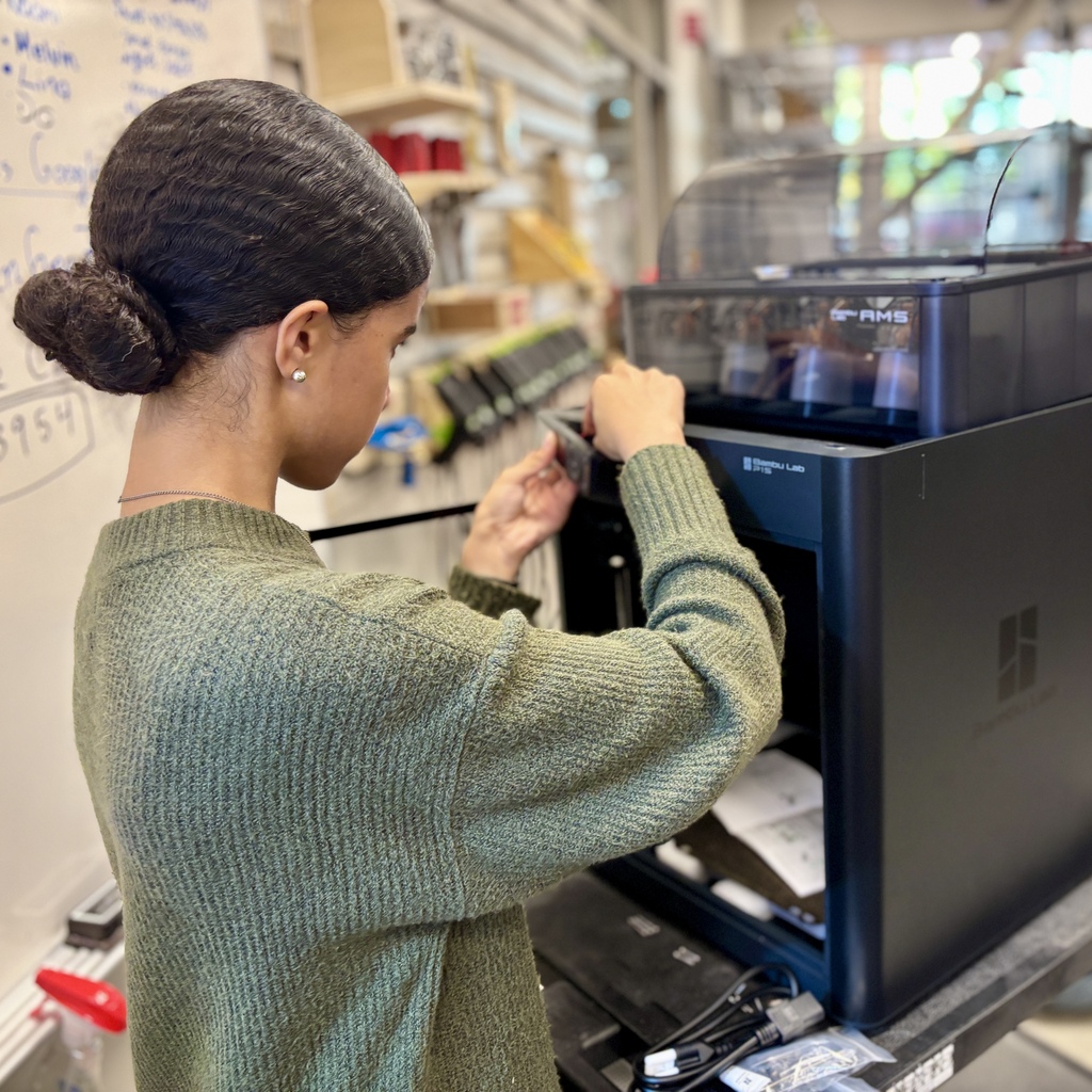 A student wearing a green sweater operates a large modern 3D printer in a workshop.