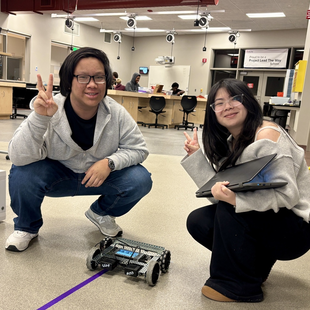 Two smiling students squat in a classroom next to a small robot on the floor, flashing peace signs.