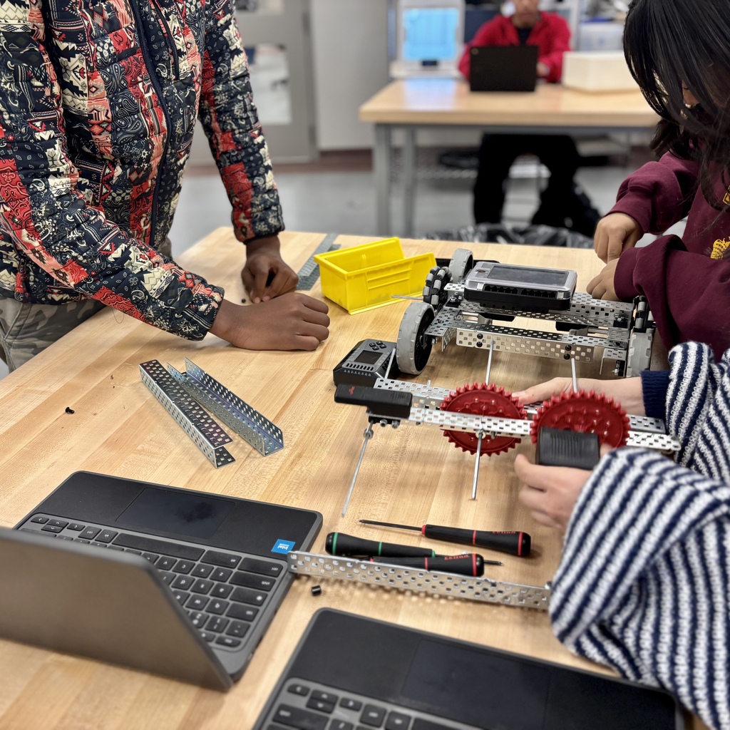 Students assemble a metal robot chassis on a wooden table with laptops.