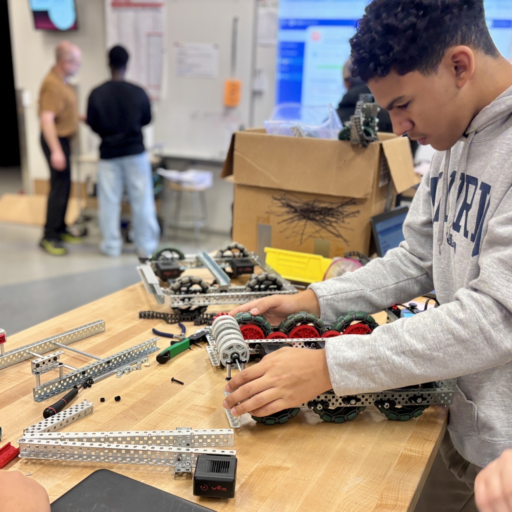 A student in a grey hoodie meticulously assembles a robotics project on a wooden workbench in a classroom.