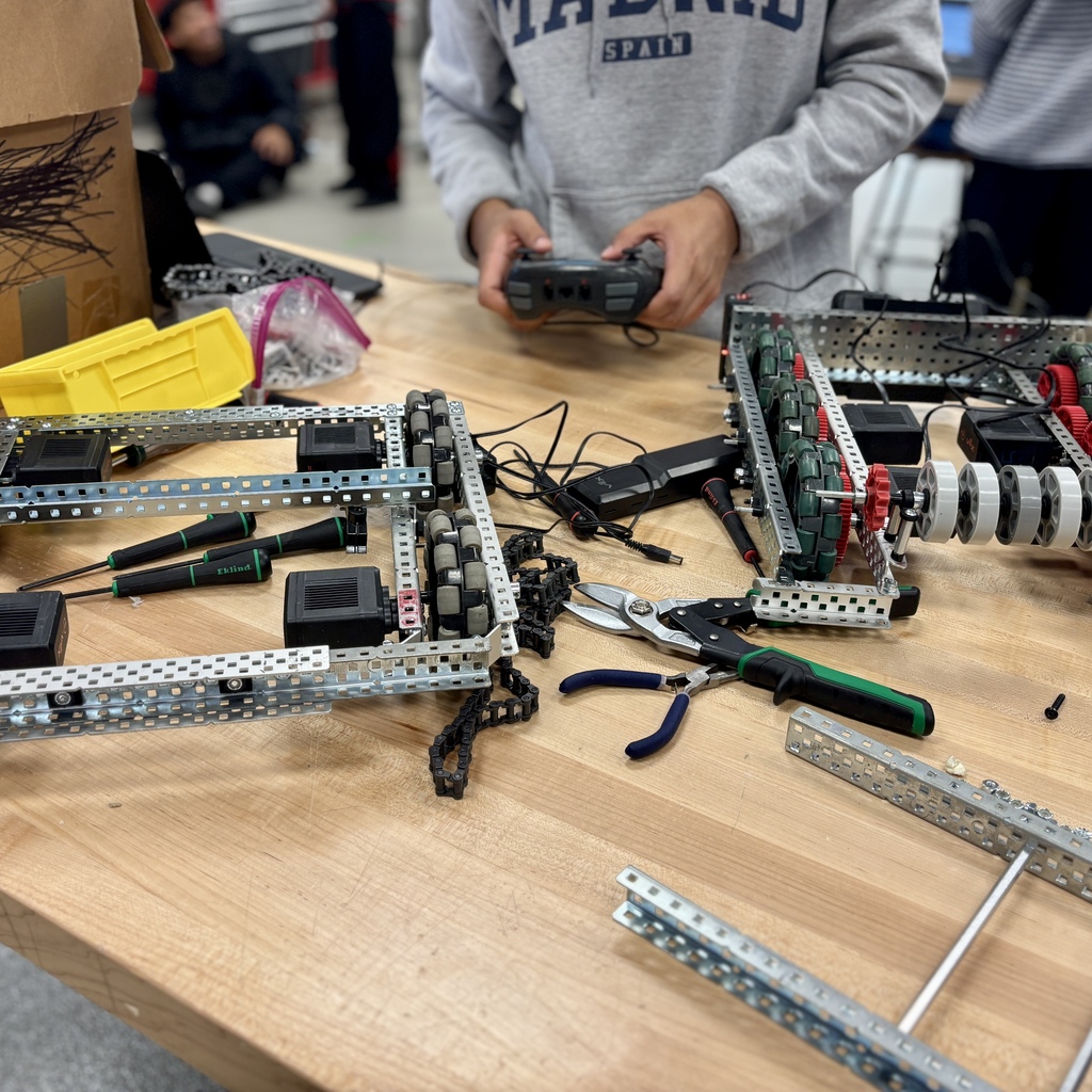 A student in a grey hoodie holds a remote, testing a metal robot chassis on a wooden workshop table.