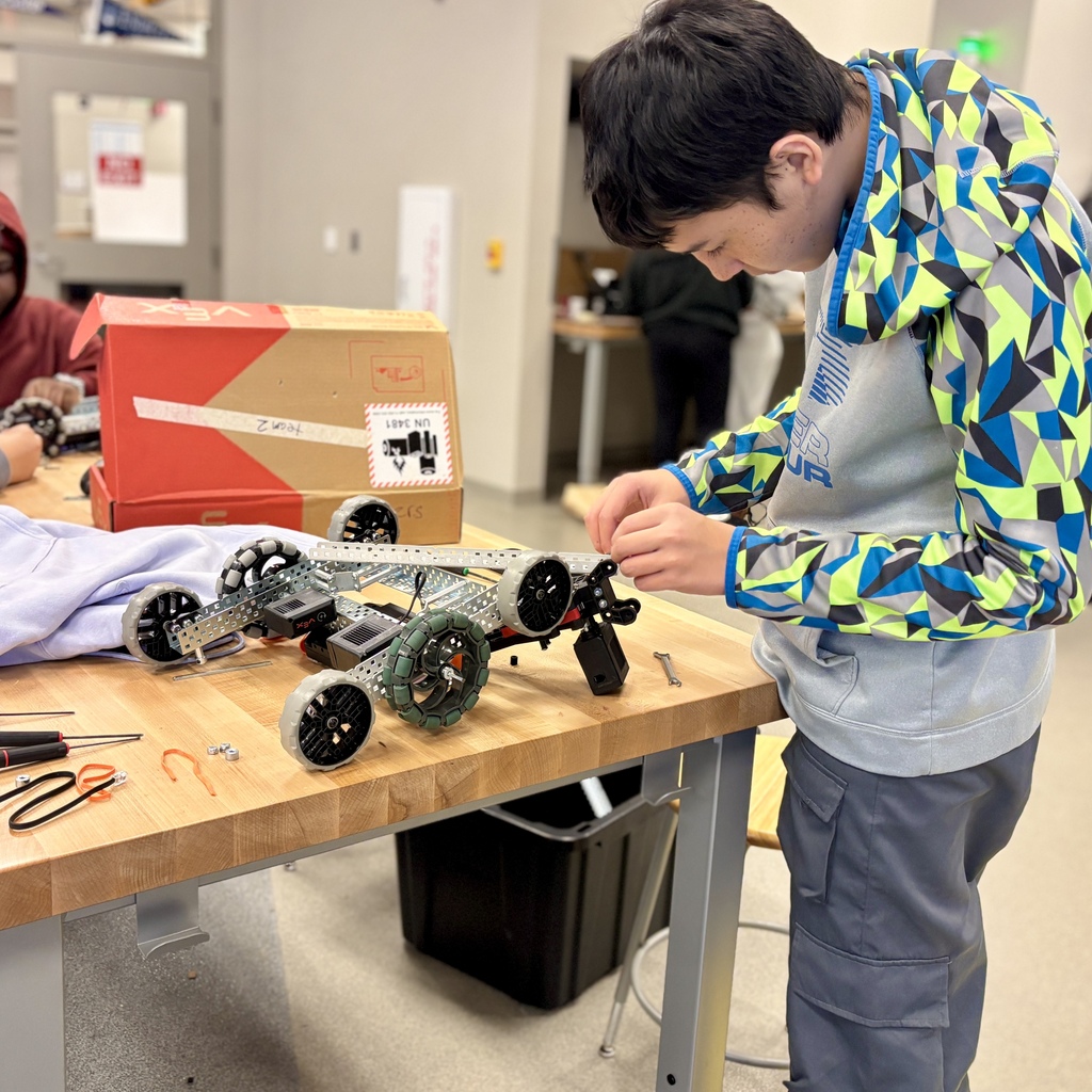 A student wearing a blue and green patterned hoodie carefully assembles a metal robot on a workbench.