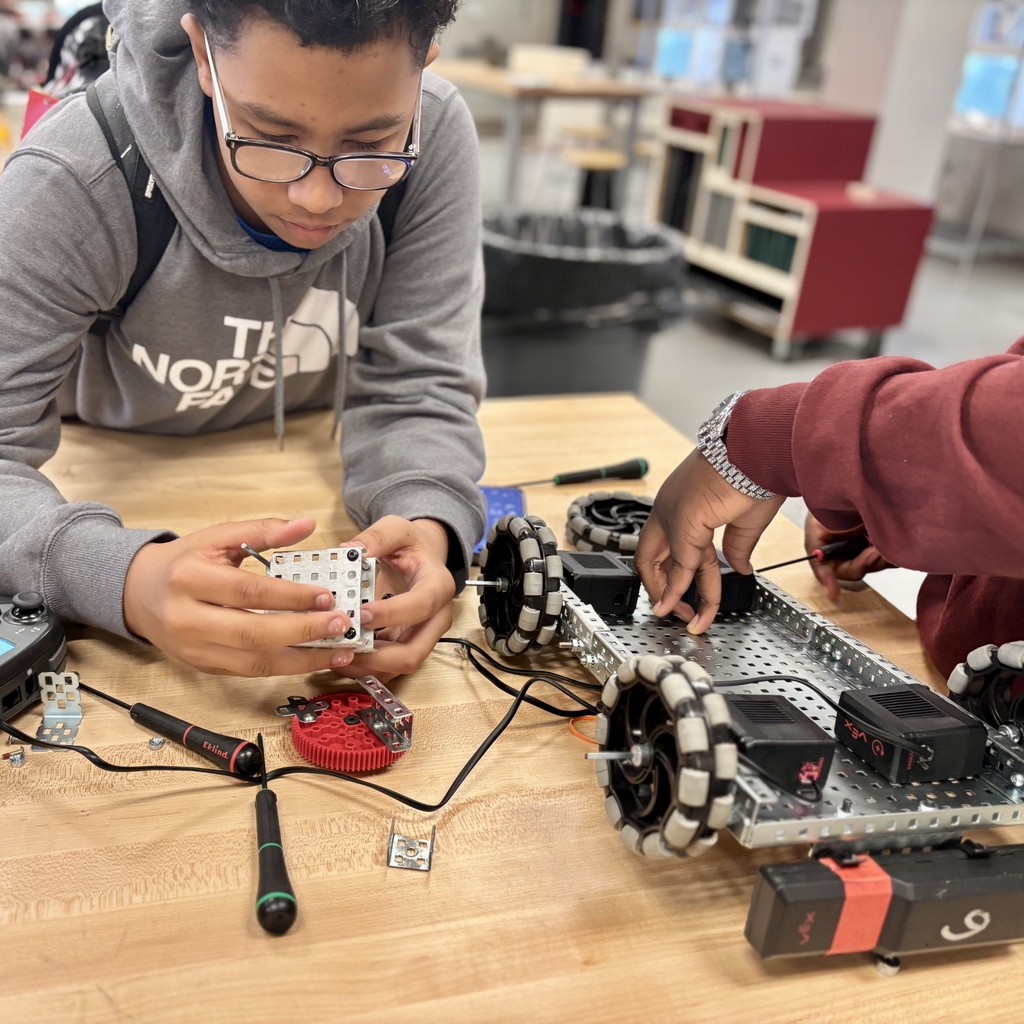 A student wearing glasses and a grey hoodie carefully assembles a small metal piece for a robotics project.