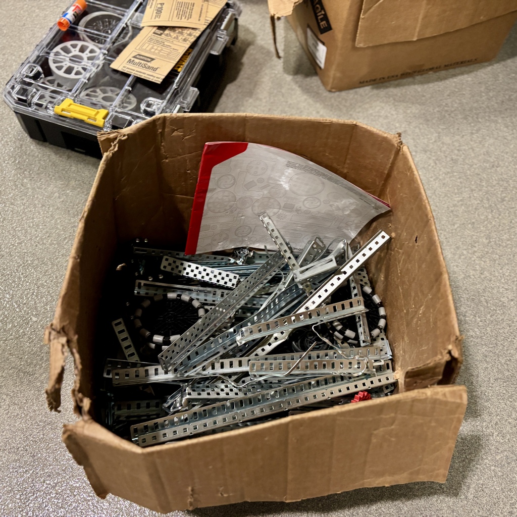 A cardboard box filled with metal construction parts and a manual rests on a grey floor.