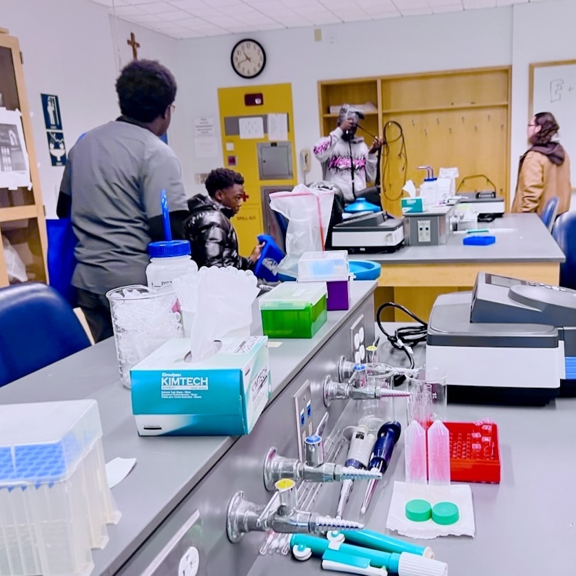 Students work in a brightly lit science lab filled with equipment, chemicals, and glassware on the counters.