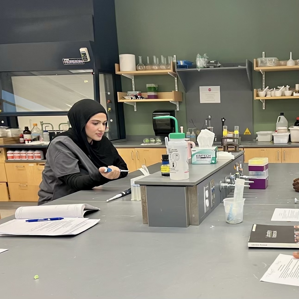A student wearing a black hijab and gray lab coat sits at a laboratory bench, holding a pipette.
