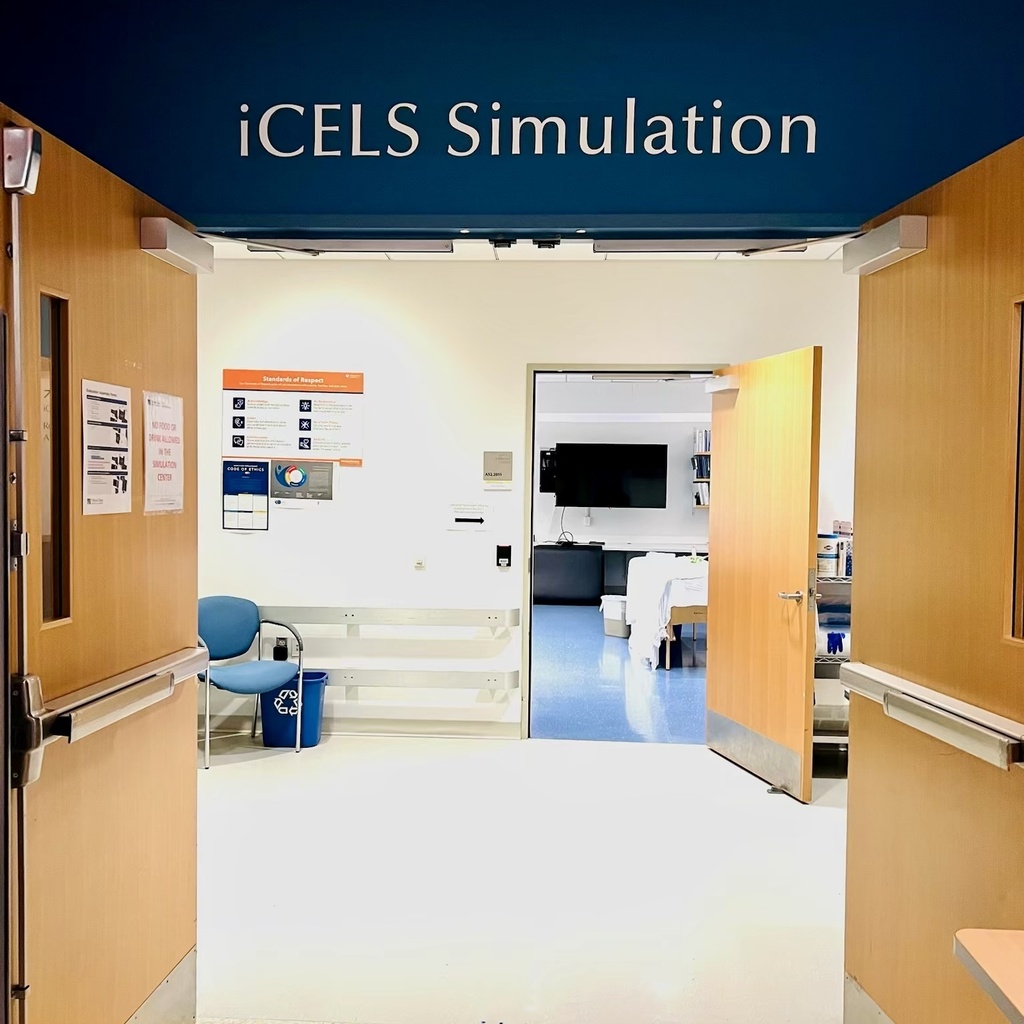 A view through open wooden doors into the iCELS Simulation room, featuring white walls, a blue chair, and a view into an adjacent medical training space.