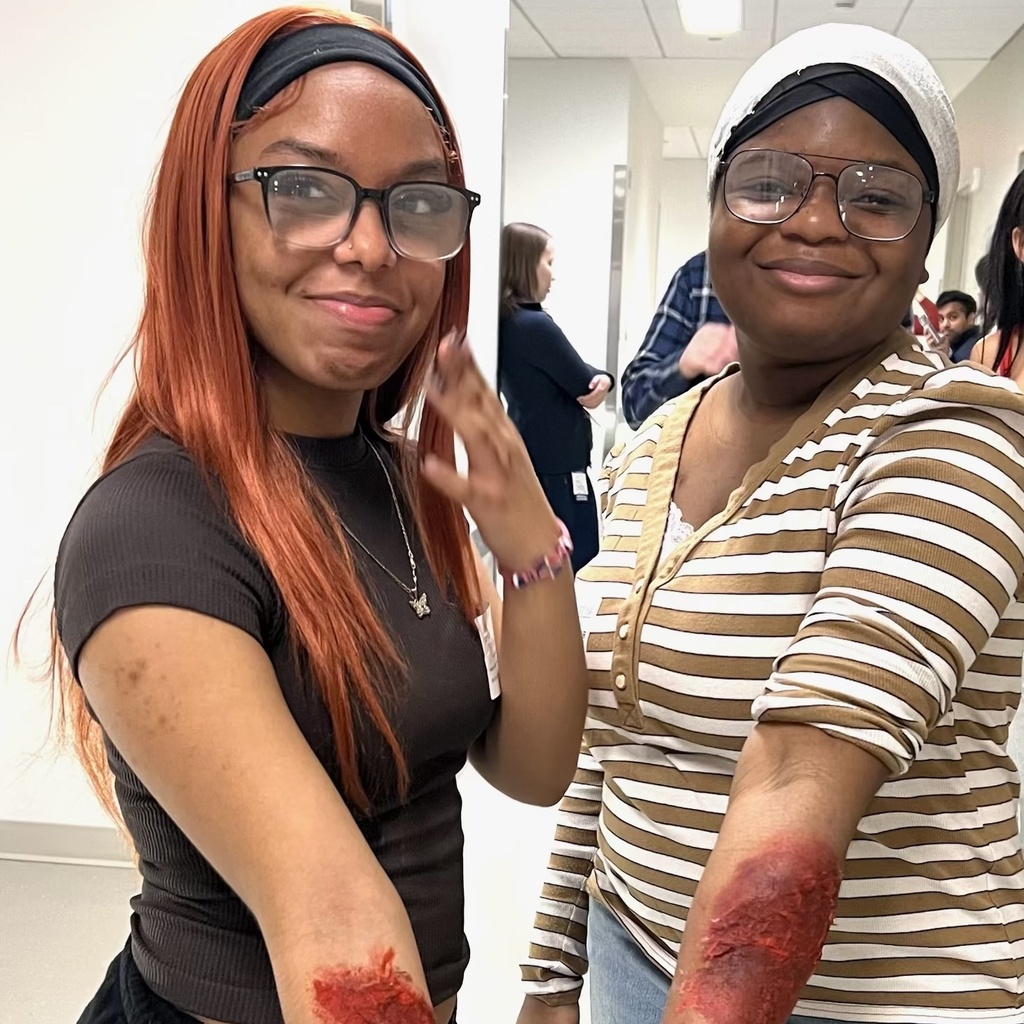 Two smiling young women proudly display theatrical makeup simulating injuries on their forearms indoors.