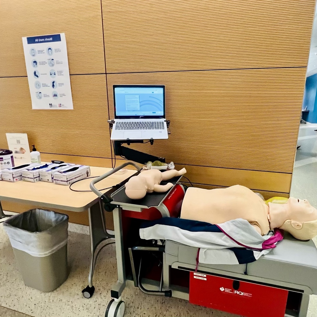 A medical training station features an adult CPR manikin and an infant manikin on a cart with an open laptop.