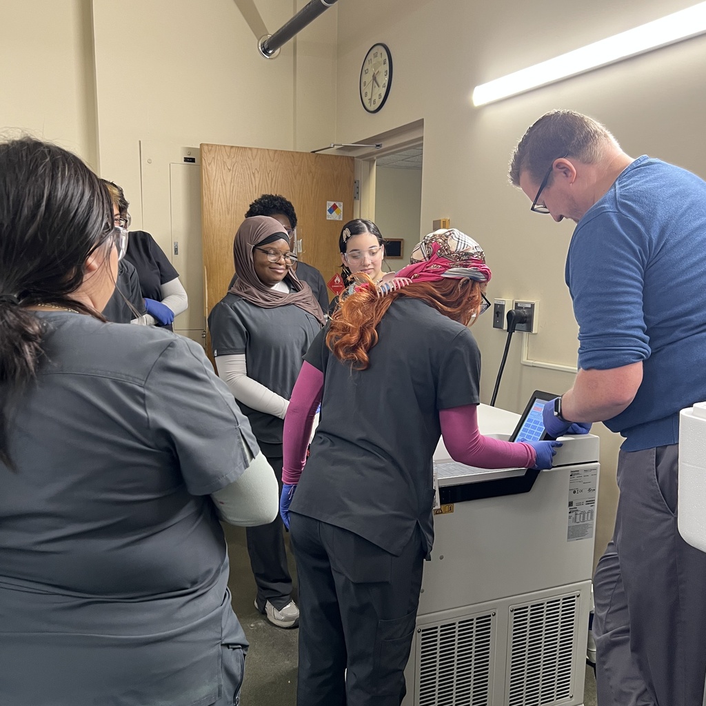A group of students in medical scrubs observe an instructor operating a lab machine.
