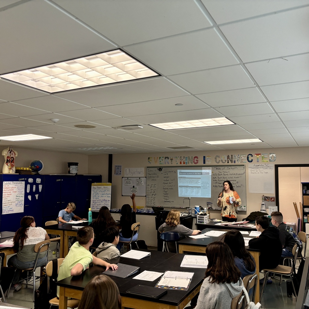 A teacher stands by a projector screen in a bright, organized classroom while students sit at lab tables, engaged in a lesson.