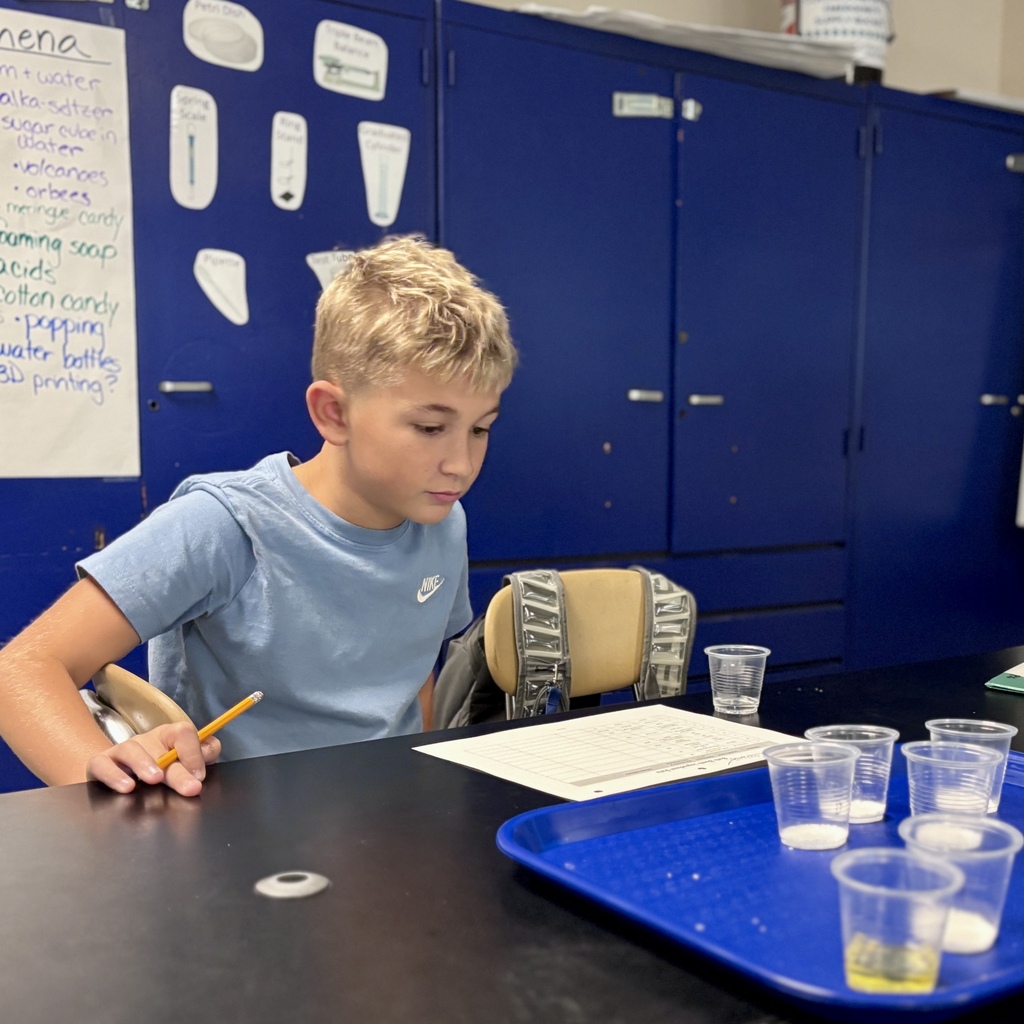 A young student sits at a science lab table, holding a pencil and looking at a worksheet.