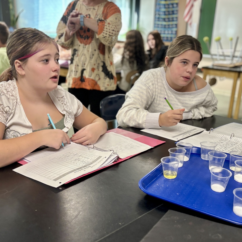 Two students sit at a table with open binders and a blue tray holding small cups during a class.