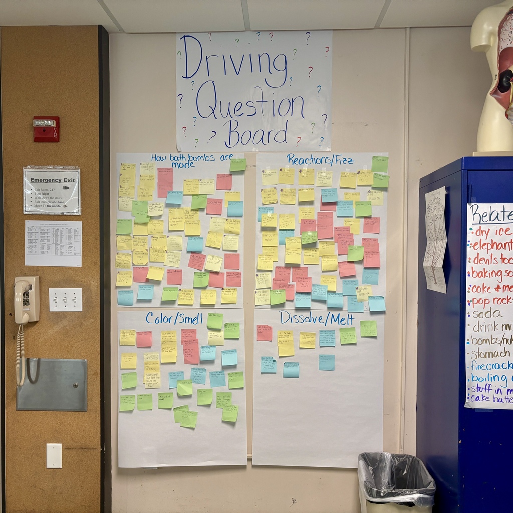 A "Driving Question Board" in a classroom, covered in colorful sticky notes with handwritten questions about bath bombs.