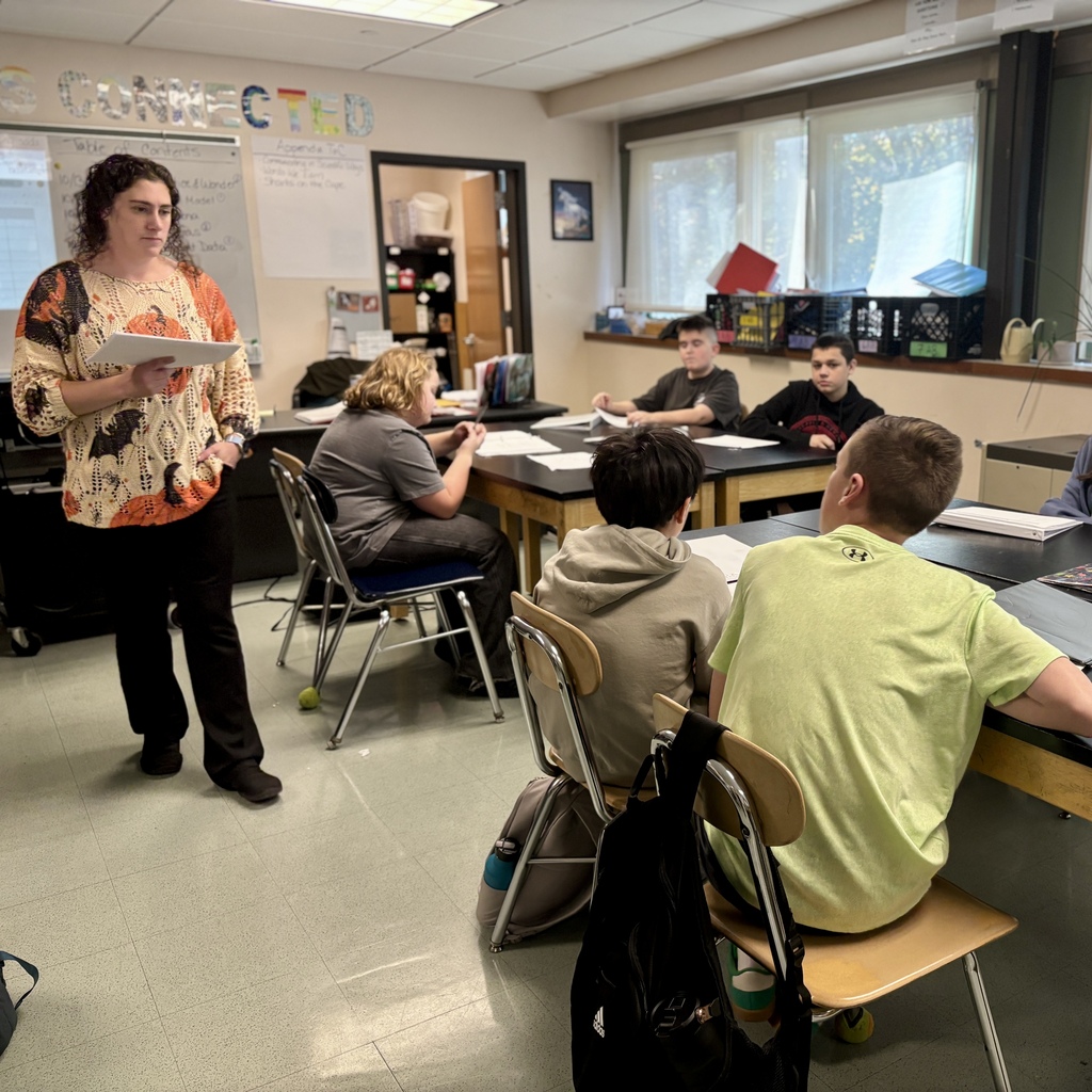 A teacher holds papers while standing at the front of a brightly lit classroom with students seated at desks.