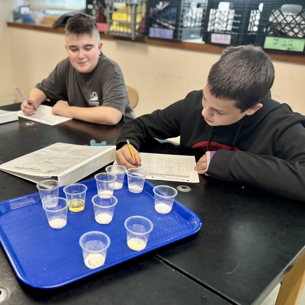 Two boys sit at a black table in a classroom, focused on writing answers on paper.
