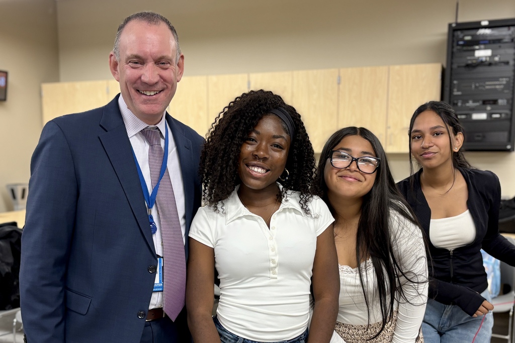 Superintendent Brian Allen smiles as he poses with three students indoors, wearing a suit and tie.