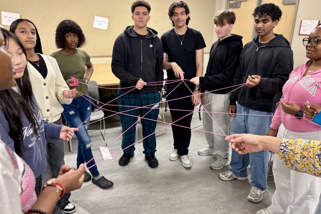 A diverse group of students stand in a circle, holding and connecting pink strings together in a classroom.