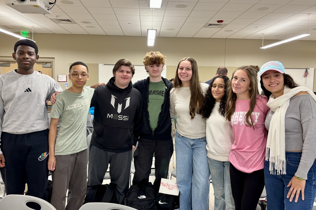 A group of eight students poses for a photo together indoors in a brightly lit room with a drop ceiling.