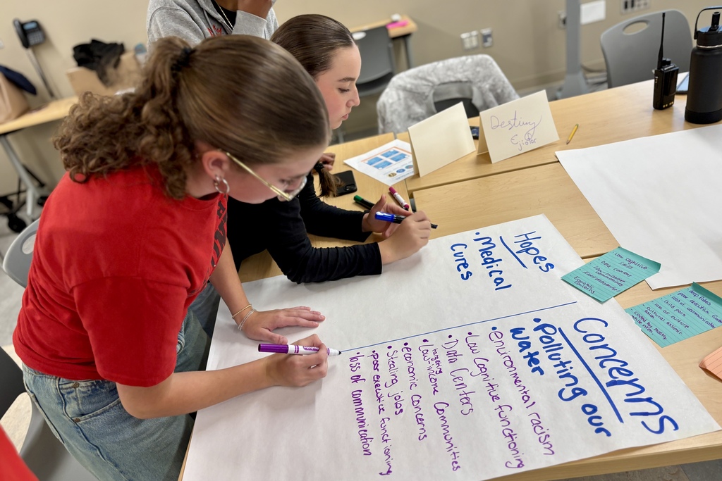 Students lean over a large sheet of paper on a table, using markers to write notes and ideas about hopes, cures, and pollution.