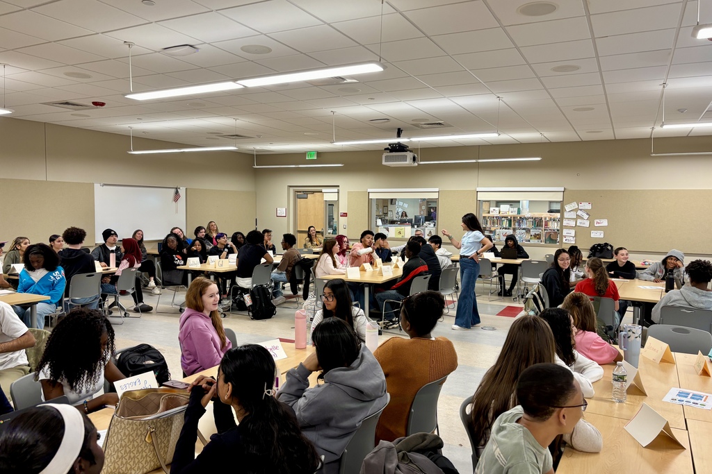 A teacher stands and speaks to a large group of high school students seated at tables in a brightly lit classroom.