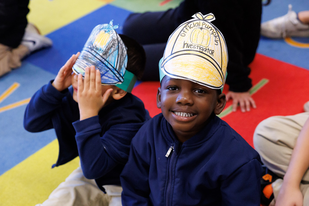 A student wearing a hat that says "official pumpkin investigator" smiles. Another is near-by, covering his eyes with his own hat.