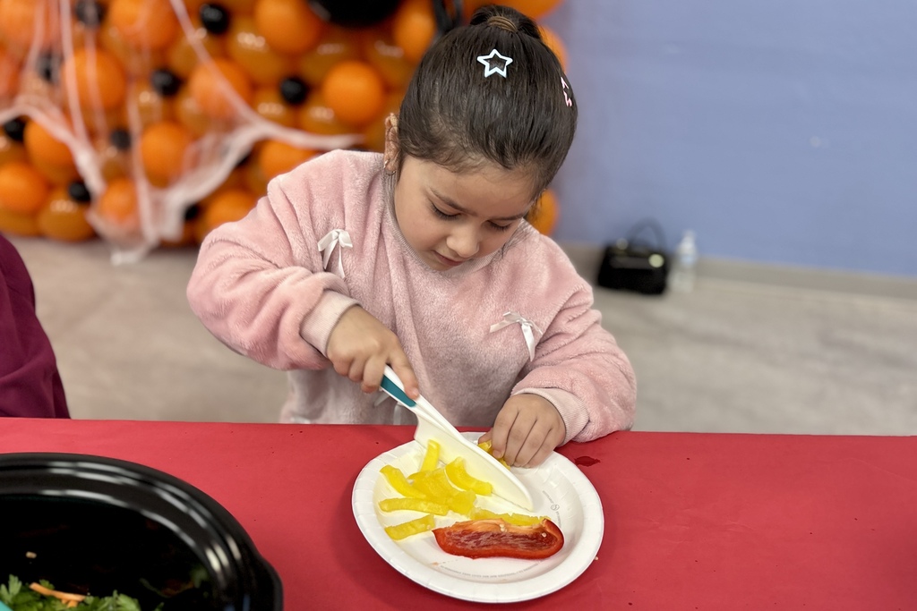 A young child in a pink sweater uses a plastic knife to cut yellow bell pepper slices on a white plate.