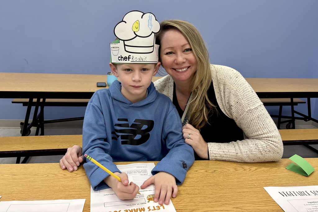 A smiling adult poses with a child wearing a paper chef hat labeled "chef RONNIE" while sitting at a table with papers.