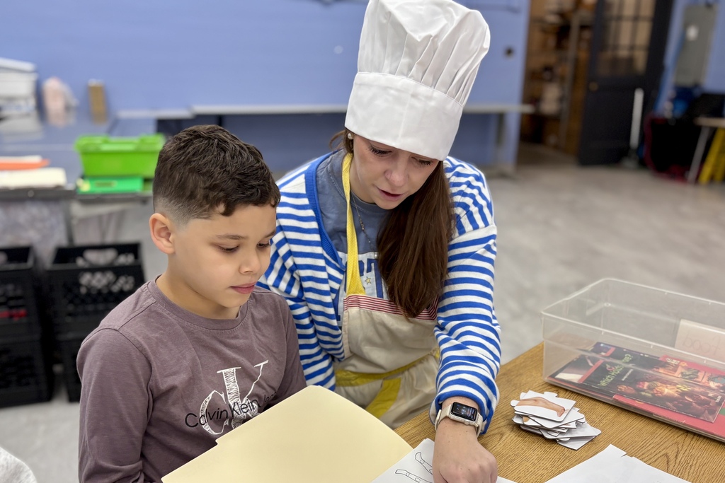 A young boy and a woman wearing a chef's hat sit together at a wooden table, looking at papers.