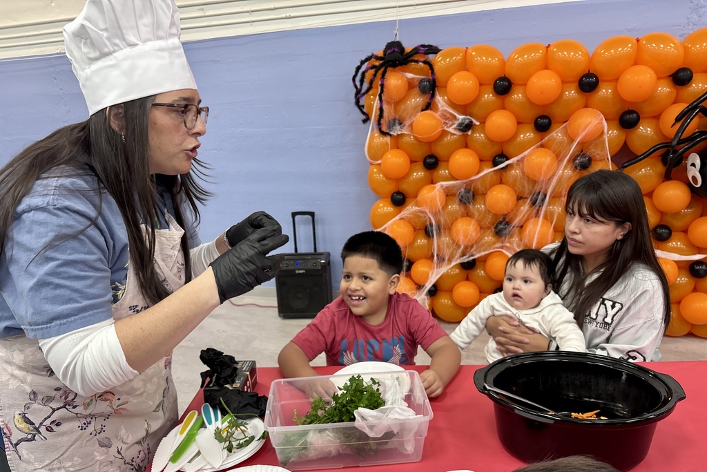A person in a chef hat and apron leads a cooking demonstration for a family at a table decorated for Halloween.