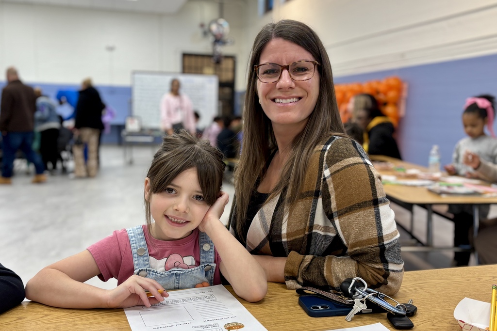 A woman and a young girl sit together at a wooden table in a large room, smiling at the camera.