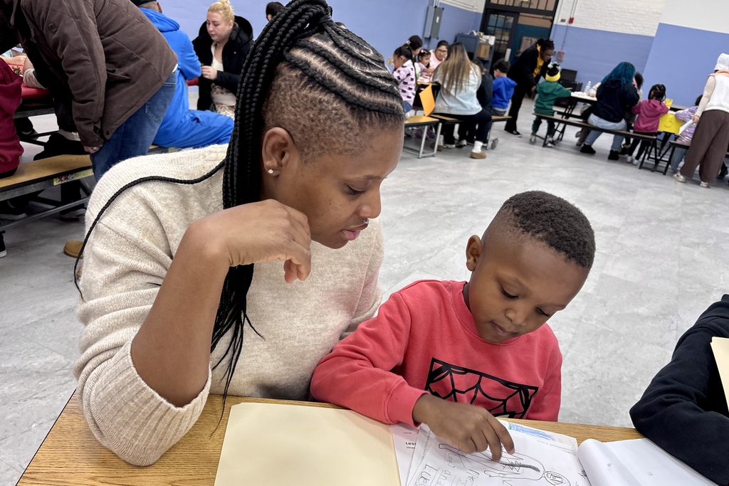 A woman with braided hair looks on as a young boy in a red shirt draws in a notebook at a table.